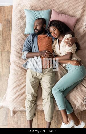 top view of dreamy african american couple embracing while lying in bed in casual clothes and looking away Stock Photo