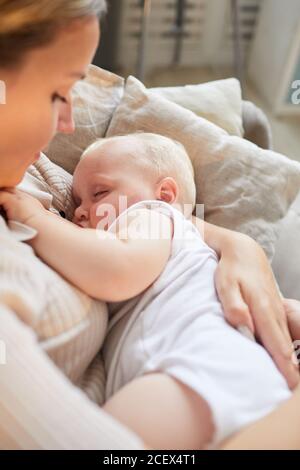Vertical high angle shot of young Caucasian mother sitting on sofa cuddling her baby son in arms Stock Photo