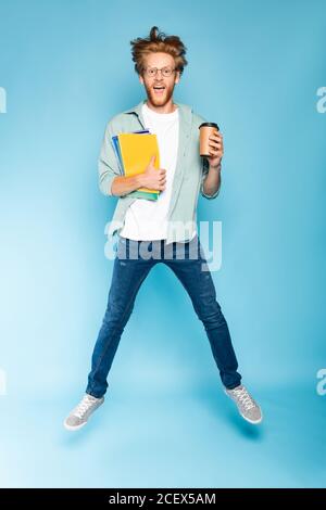 bearded student in glasses holding paper cup and notebooks while jumping on blue Stock Photo