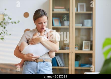 Horizontal medium shot of young Caucasian mother standing in living room cuddling her baby son in arms Stock Photo