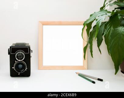 Mock up wooden frame, old camera, plant and pencils. Interior home square poster mockup with wood frame and green leaves on white wall background Stock Photo
