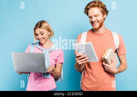 redhead student holding books and using digital tablet near blonde friend with laptop on blue Stock Photo