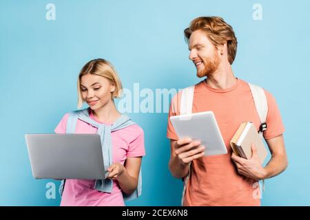 redhead student holding books and digital tablet while looking at blonde friend using laptop on blue Stock Photo