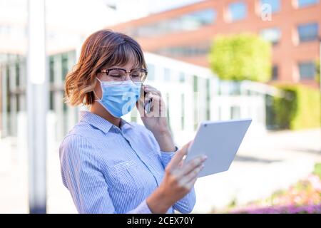 Young 20s businesswoman in mask calling and using tablet outdoors in park Stock Photo
