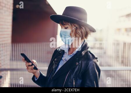 Young trendy woman in mask using smartphone outdoors in city Stock Photo