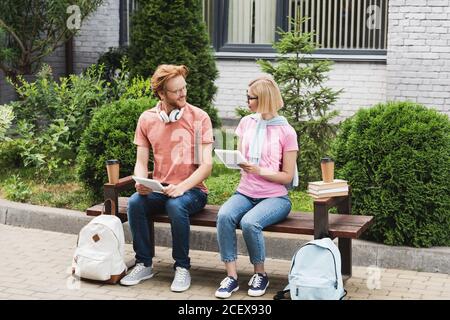 bearded and blonde students sitting on bench near books, paper cups and backpacks while holding digital tablets Stock Photo