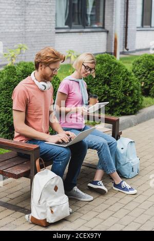 young students sitting on bench and using gadgets while online study outside Stock Photo