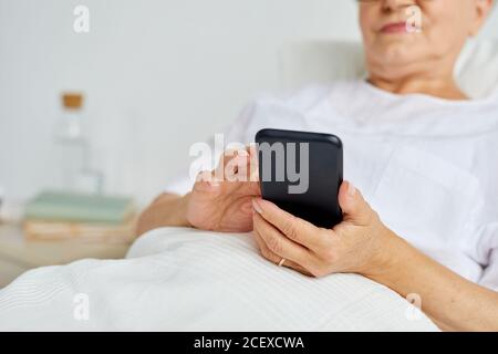 Modern aged woman wearing white clothes relaxing on bed in hospital ward texting messages using her smartphone Stock Photo
