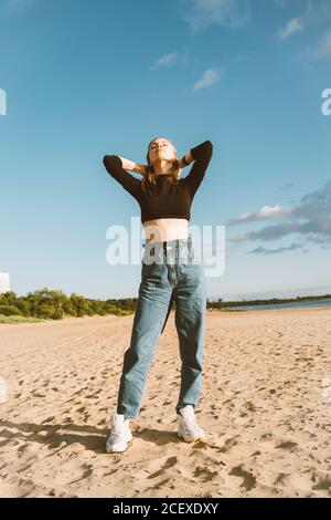 Full length beautiful woman standing on beach at sunset in evening. Pretty female with long brown hair in casual clothes, hands on head, looking up Stock Photo