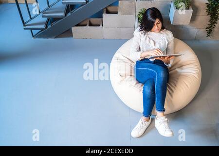 Top view full length of focused young female designer in casual clothes sitting on comfortable pouf and using graphic tablet while working in contemporary creative workspace