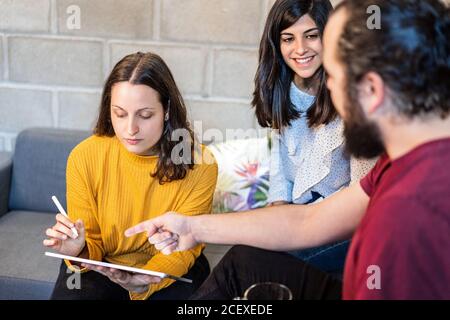 Group of creative millennial designers drinking coffee and discussing design for new project on graphic tablet while gathering in modern loft coworking space Stock Photo