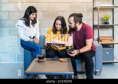 Group of creative millennial designers drinking coffee and discussing design for new project on graphic tablet while gathering in modern loft coworking space Stock Photo