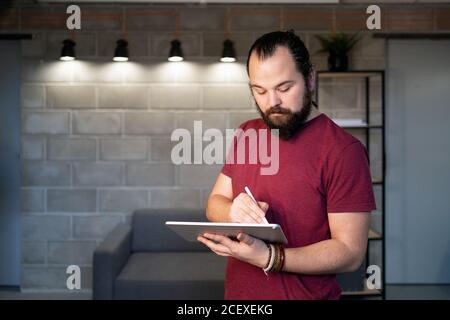 Focused young bearded male freelancer in casual outfit standing using tablet while working on project in contemporary loft workspace Stock Photo