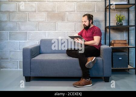 Full body of focused young bearded male freelancer in casual outfit sitting on sofa and using tablet while working on project in contemporary loft workspace Stock Photo