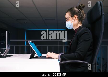 Side view of businessWoman specialist sitting at table and typing on keyboard on tablet with empty white screen while working at workplace Stock Photo