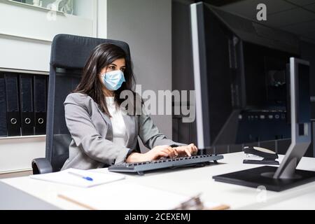 Confident young female specialist in formal suit and protective mask sitting at desk and using computer while working in modern workspace Stock Photo