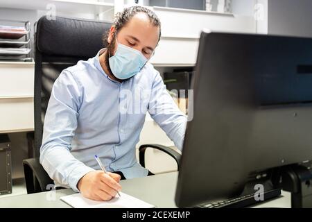 Focused young bearded male manager in formal blue shirt and protective mask making notes in notebook while sitting in chair in front of computer monitor in modern workplace Stock Photo