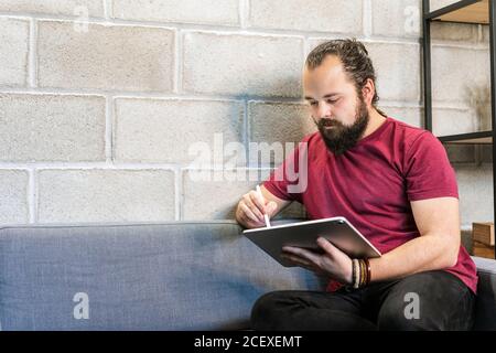 Focused young bearded male freelancer in casual outfit sitting on sofa and using tablet while working on project in contemporary loft workspace Stock Photo