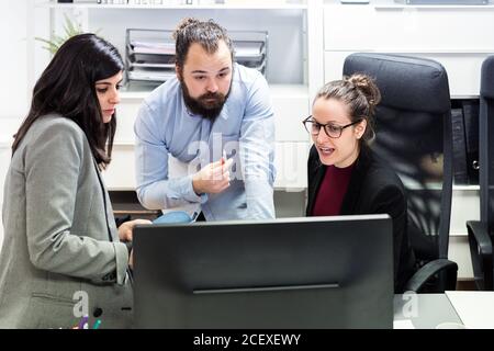 Serious young businessman and female colleagues gathering around computer and discussing business data while working together in contemporary workspace Stock Photo