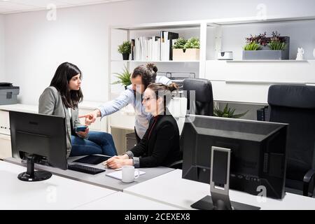 Serious young businessman and female colleagues gathering around computer and discussing business data while working together in contemporary workspace Stock Photo