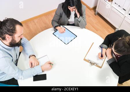 From above serious young man and women in formal clothes sitting at table and making notes while working together on business strategy in contemporary coworking office Stock Photo