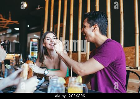 Cheerful young man giving snack to laughing girlfriend while having lunch with friends in cafeteria Stock Photo
