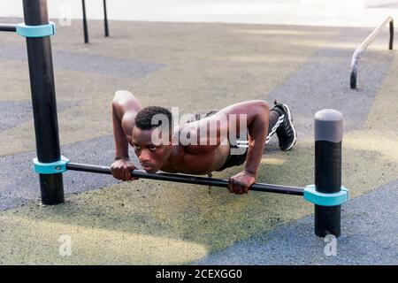 Concentrated black sportsman doing push ups wit metal crossbar during active workout and looking forward Stock Photo
