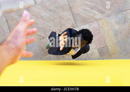 From above of crop person giving helping hand for African American male standing on stone ground near yellow building Stock Photo