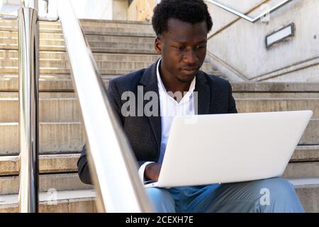 Serious African American male entrepreneur sitting on stone stairs in city and working on remote project while using modern netbook Stock Photo