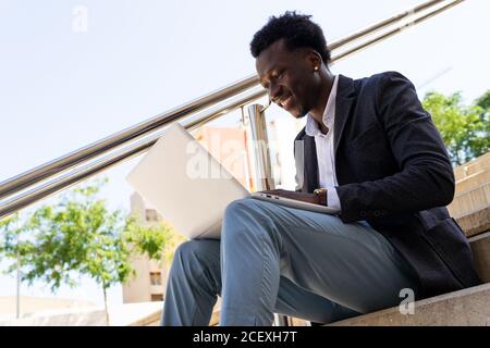 Serious African American male entrepreneur sitting on stone stairs in city and working on remote project while using modern netbook Stock Photo