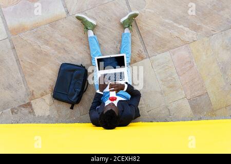 Top view of unrecognizable African American businessman in stylish clothes with briefcase drinking coffee and using laptop while sitting near wall on city street Stock Photo