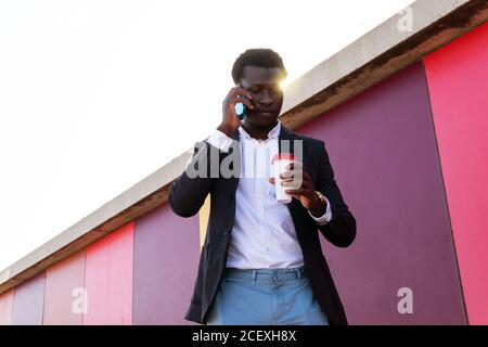 Low angle of serious young African American businessman in trendy clothes holding disposable cup of coffee and discussing business news via smartphone while standing near colorful wall on street Stock Photo