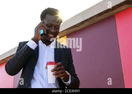 Low angle of serious young African American businessman in trendy clothes holding disposable cup of coffee and discussing business news via smartphone while standing near colorful wall on street Stock Photo