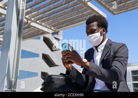 Low angle of busy African American male manager in protective mask sitting in city and browsing cellphone while working remotely on project during COVID 19 outbreak Stock Photo