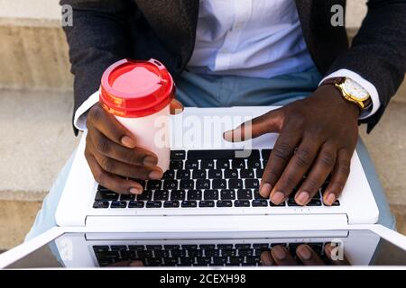 Cropped unrecognizable African American businessman in formal suit drinking takeaway coffee while sitting on city street and browsing laptop Stock Photo