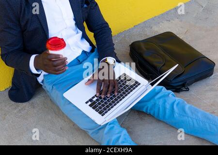 Cropped unrecognizable African American businessman in stylish clothes with briefcase drinking coffee and using laptop while sitting near wall on city street Stock Photo