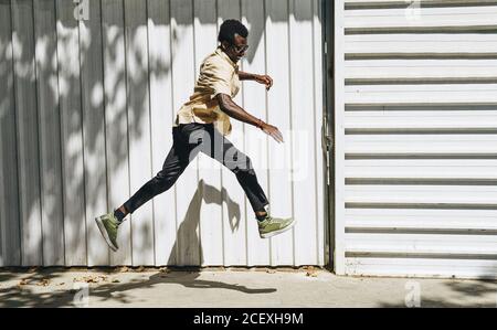Side view of smiling African American male in stylish wear in moment of jumping near building on sunny day during city stroll Stock Photo