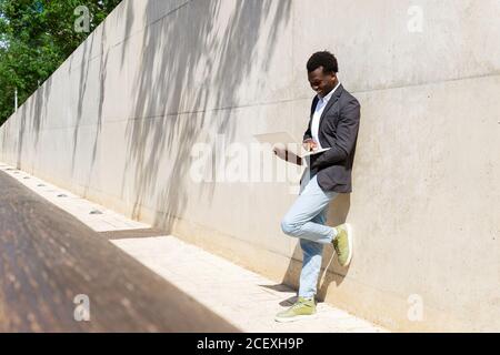 Focused African American male executive manager standing against concrete wall in the street with laptop Stock Photo