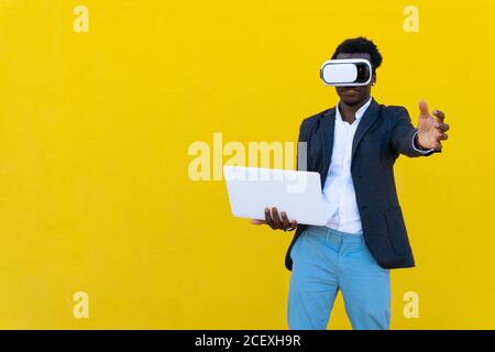 African American male entrepreneur standing with laptop and experiencing virtual reality while using modern VR headset on yellow background in city Stock Photo