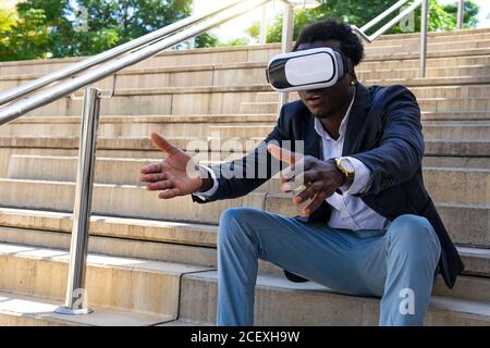 Curious ethnic businessman sitting on stone stairs in VR goggles and experiencing virtual reality with outstretched arms Stock Photo