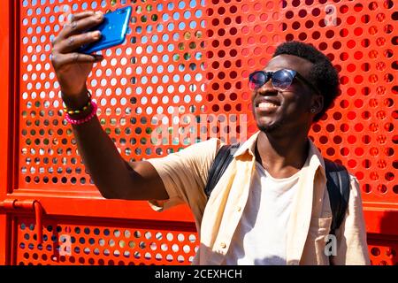 Content black male in summer wear and sunglasses standing near red metal wall and taking photo on mobile phone camera during city stroll Stock Photo