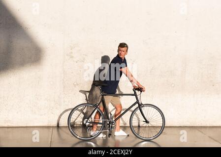 Side view of casual dressed young man walking with his urban bicycle near concrete wall Stock Photo