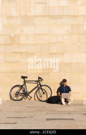 Young entrepreneur working outdoors on his laptop with his bicycle leaning on concrete wall Stock Photo