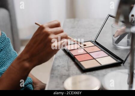 Side view of crop anonymous female taking eye shadow from palette with makeup brush during beauty procedure at home Stock Photo