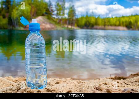 A blue plastic bottle with an open lid stands on a sandy beach against the backdrop of a wild lake. Background. Stock Photo