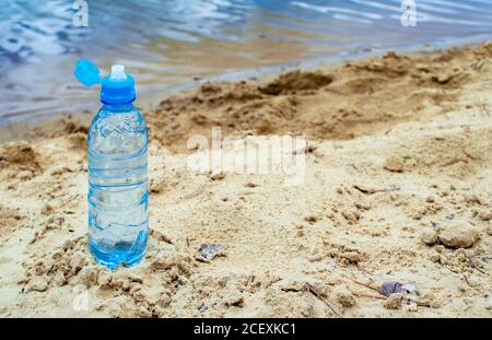 A blue plastic bottle with an open lid stands on a sandy beach against the background of lake water. Background. Stock Photo