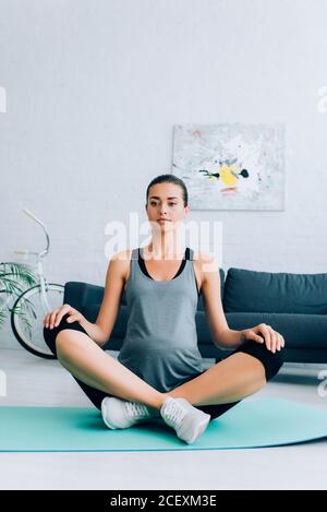Pregnant sportswoman sitting with crossed legs in yoga pose on fitness mat at home Stock Photo