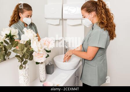 Side view of young female beautician doctor in protective mask washing hands while preparing for treatment procedure in modern office Stock Photo