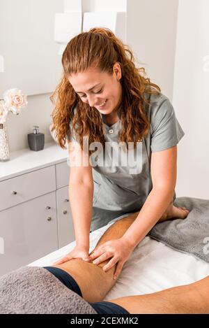 Smiling young professional masseuse doing anti cellulite massage on legs of anonymous patient while working in modern beauty salon Stock Photo