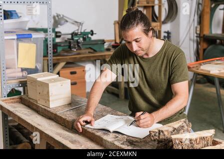Concentrated male carpenter standing at workbench and taking notes in notebook while working in garage Stock Photo
